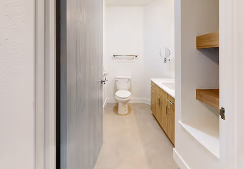View into a clean, modern bathroom featuring a white toilet centered against the back wall, a wooden vanity with a white countertop and a round mirror on the right, beige hexagonal floor tiles, and built-in wooden shelves on the right wall.