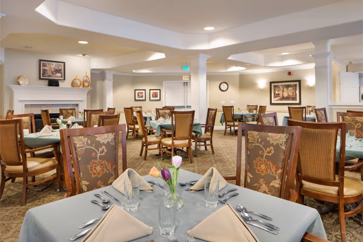 A dining room in a senior living facility with multiple tables covered in light blue tablecloths, each set with beige folded napkins, glasses, and silverware. The chairs have wooden frames with floral upholstery. The room has beige walls decorated with framed artwork, a fireplace, and a clock on the wall. The lighting is warm and recessed in the ceiling.
