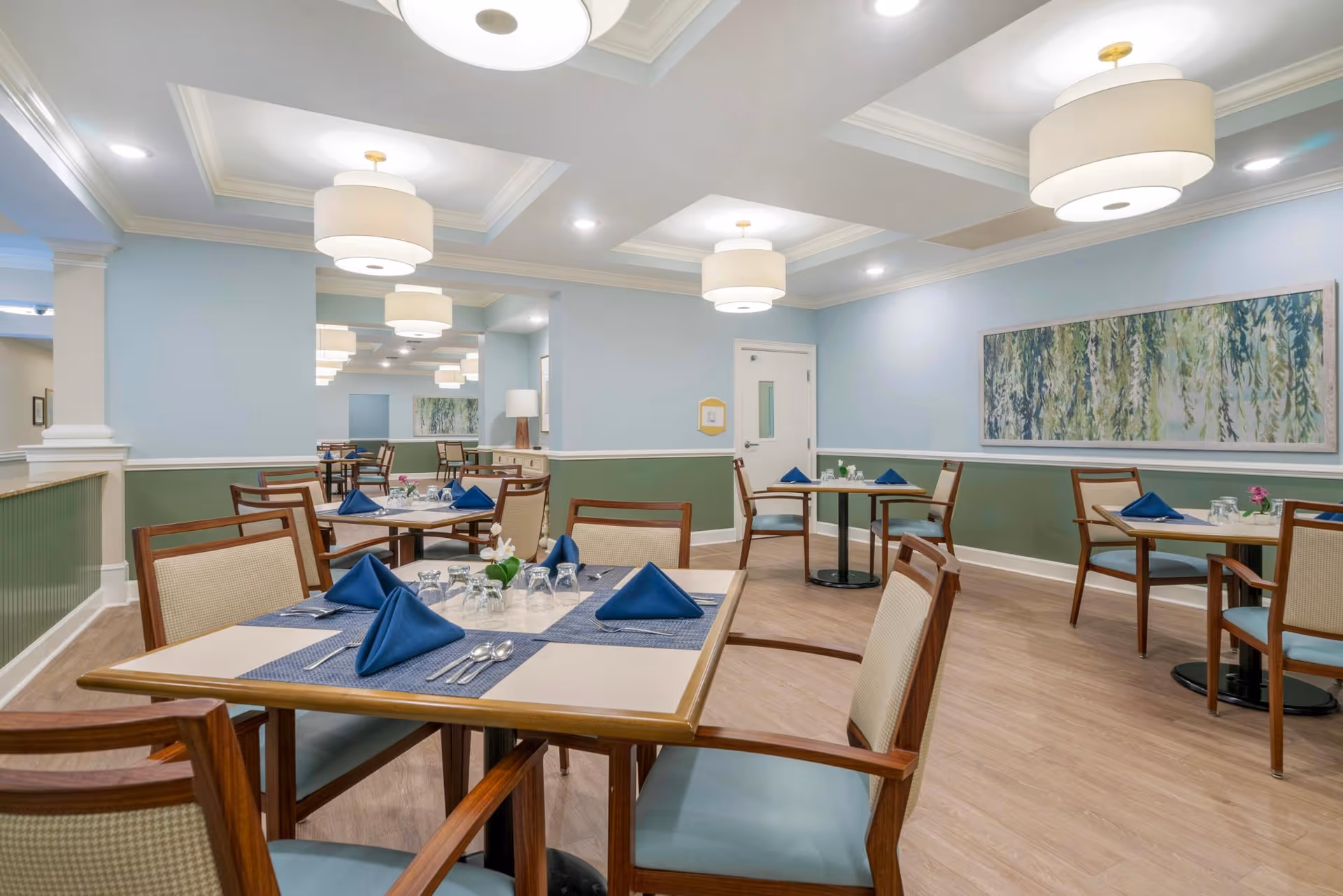 A dining room in a senior living facility with several tables set with blue napkins, glasses, and silverware. The room features light blue and green walls, wooden chairs with cushioned seats, modern ceiling lights, and a large abstract painting on the wall.