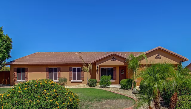 Single-story house with a brown tiled roof and beige exterior walls, surrounded by green shrubs, palm trees, and a well-maintained lawn under a clear blue sky.