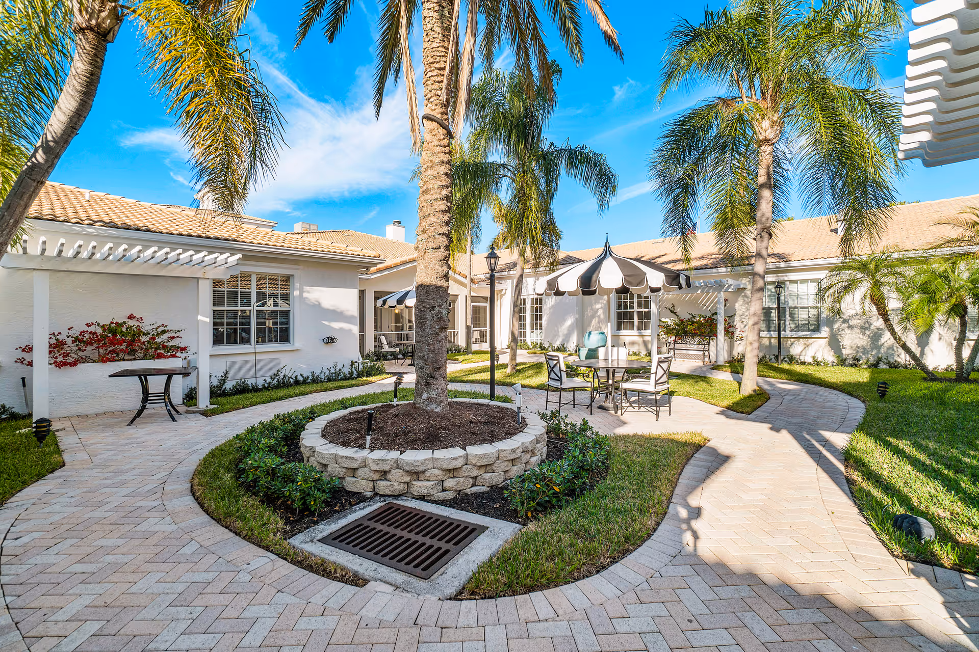 Outdoor courtyard area at Tequesta Terrace featuring a paved walkway winding around a circular raised garden bed with palm trees. There are patio tables with black and white striped umbrellas, chairs, and lush green landscaping under a bright blue sky.