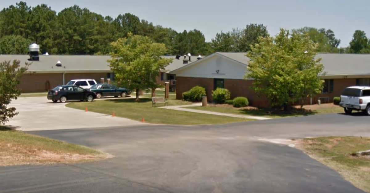 Exterior view of a single-story brick building with a gray roof, surrounded by trees and parked cars in the driveway, under a clear sky.