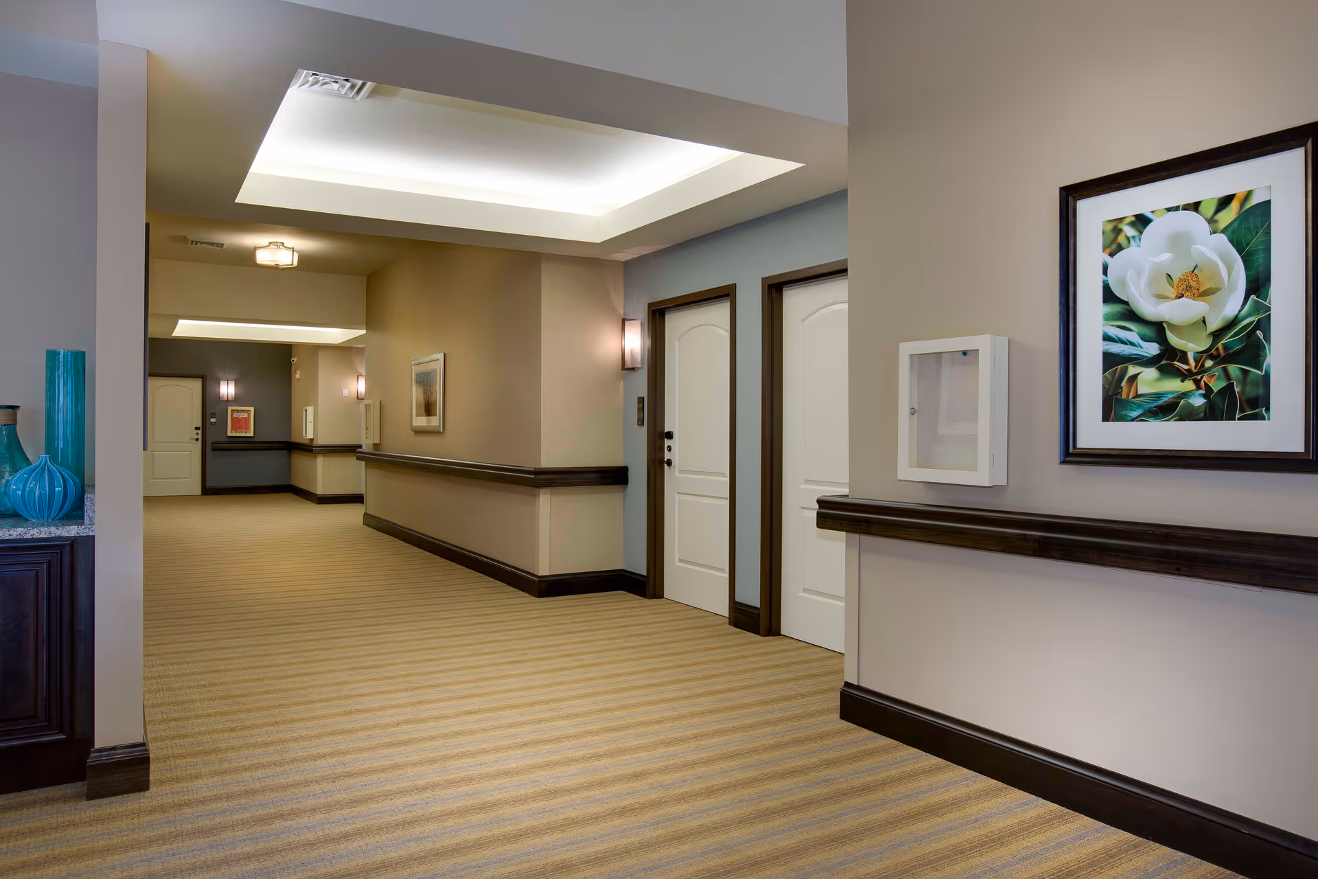 A well-lit hallway in a senior living facility with beige walls and striped carpet. The hallway features white doors with dark trim, a framed floral painting on the wall, decorative vases on a dark wooden cabinet, and modern ceiling lights.