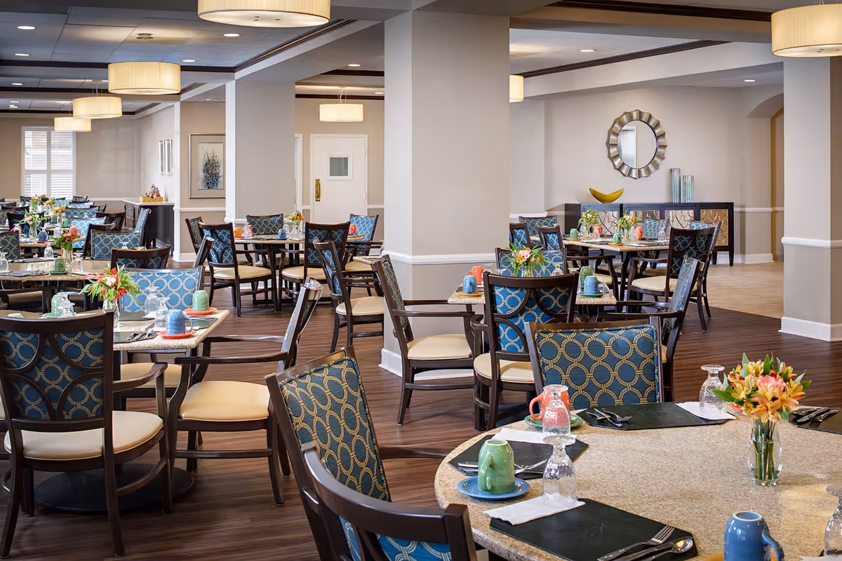 A spacious and well-lit dining room in a senior living facility with multiple round tables set with colorful mugs, glasses, utensils, and small flower arrangements. The room features wooden flooring, patterned chairs with blue and gold upholstery, beige walls, and modern ceiling lights. A decorative mirror and sideboard are visible in the background.