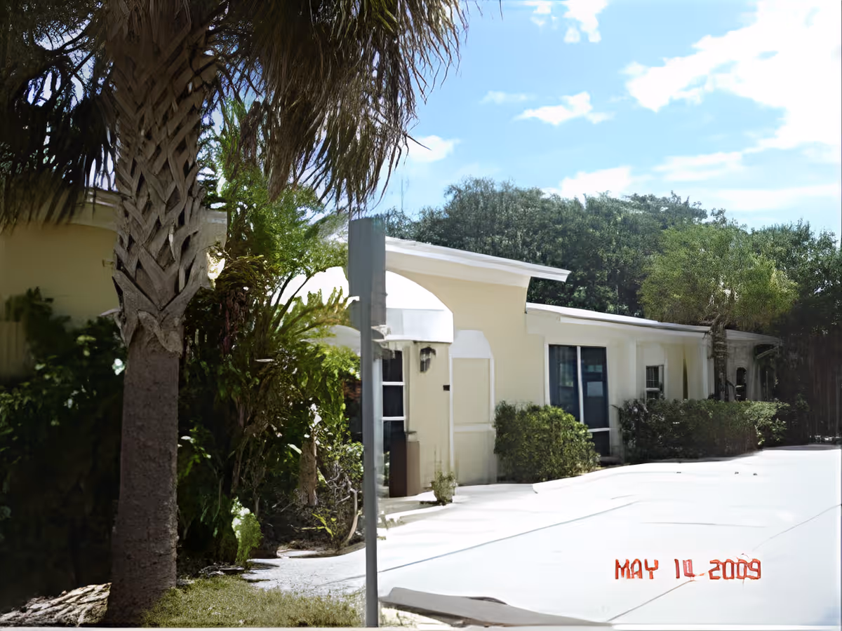 Single-story light-colored building with a palm tree, shrubs and a driveway under a sunny sky.
