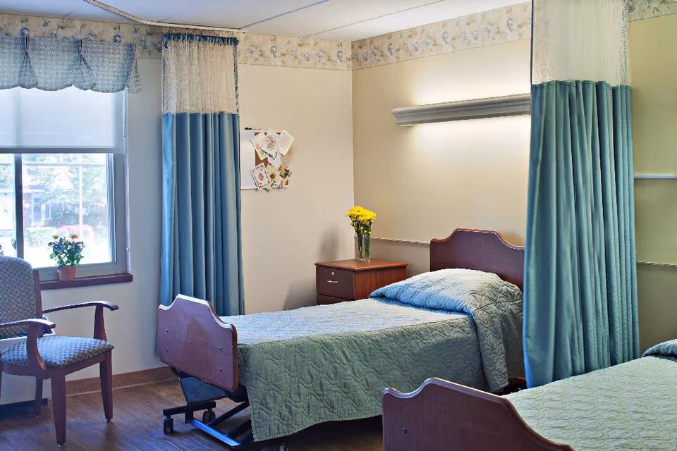 A senior living facility bedroom with two single beds covered in light blue quilts. There is a wooden nightstand between the beds with a vase of yellow flowers on it. A window with blue curtains and a valance lets in natural light, and a cushioned wooden chair is placed near the window. The room has light-colored walls with a floral border near the ceiling and a wall-mounted light fixture above one bed.
