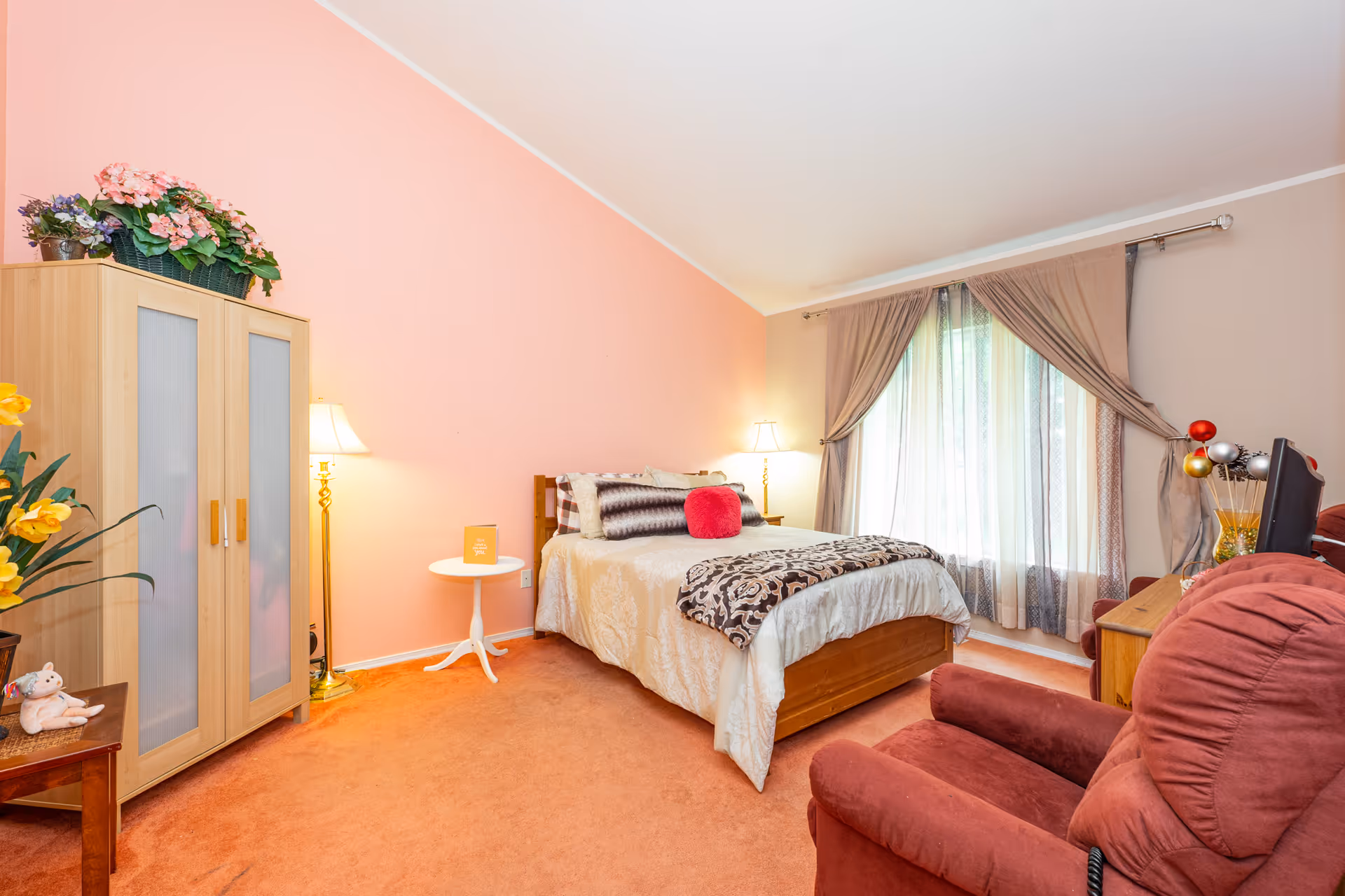 A cozy bedroom with a bed covered in beige and patterned bedding, a red throw pillow, and two lamps on either side. There is a wooden wardrobe with frosted glass doors and a basket of flowers on top. A large window with beige curtains lets in natural light. In the foreground, there is a red armchair and a wooden table with decorative items. The walls are painted peach and beige, and the carpet is orange.