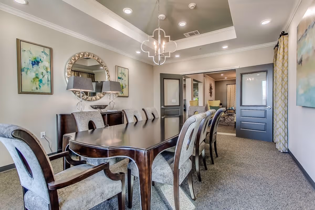 A formal dining room with a long dark wooden table surrounded by eight upholstered chairs. The room features a modern chandelier, a round decorative mirror, two table lamps on a sideboard, and abstract artwork on the walls. Double doors open to an adjacent room with additional seating.