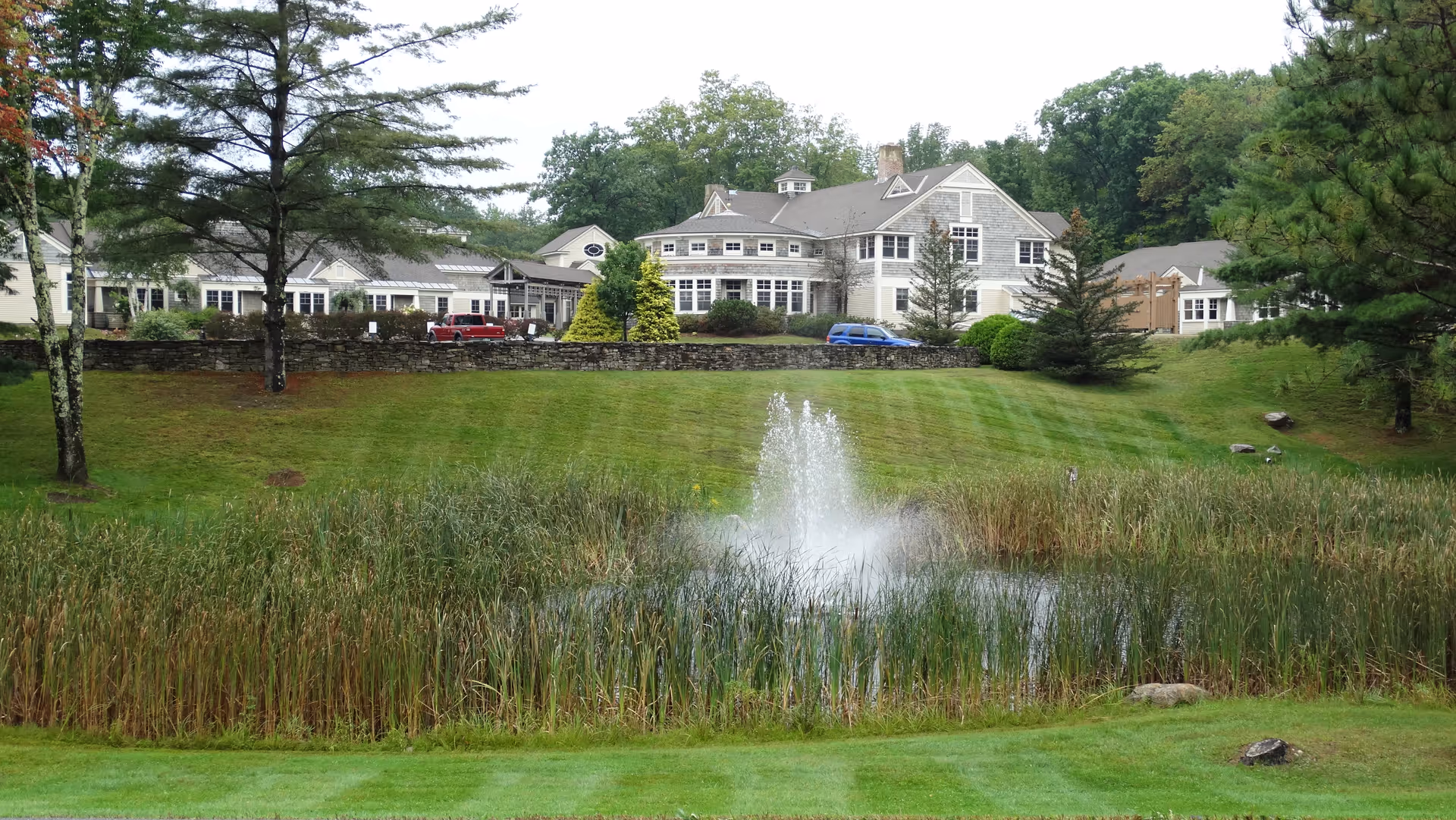 A large senior living facility building named BROMLEYMANOR is visible in the background, surrounded by trees and greenery. In the foreground, there is a pond with tall reeds and a water fountain spraying water upwards. The area is well-maintained with green grass and landscaping.