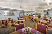 A spacious dining room with multiple wooden tables and chairs featuring purple cushions. Each table is set with placemats, napkins, and condiments. The room has a carpeted floor, light-colored walls, and ceiling lights. In the background, there is a counter area with kitchen equipment and a wall clock.