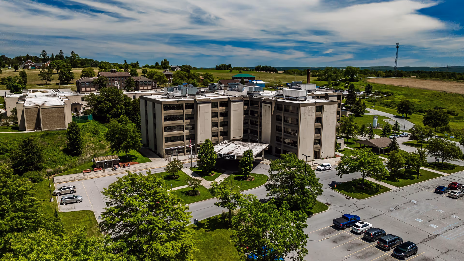 Aerial view of a multi-story health and rehab building surrounded by lawns, trees, parking lots, and open fields under a partly cloudy sky.