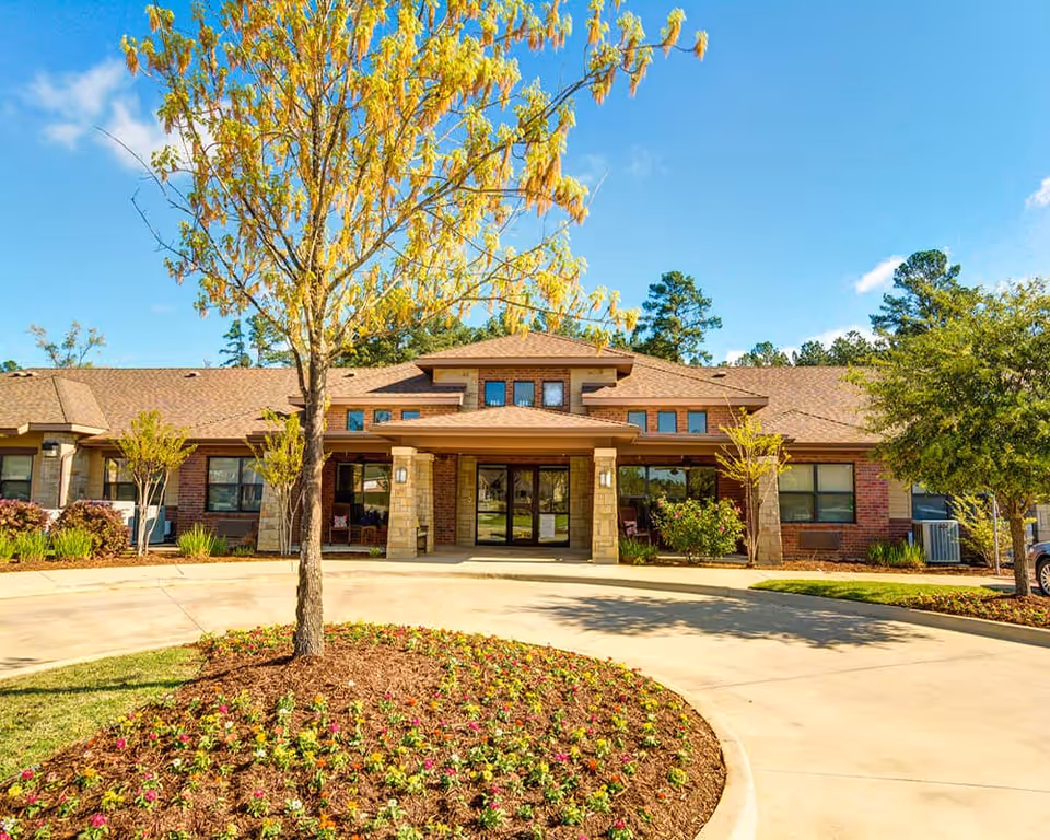 Front entrance of a single-story brick building with a covered entry, circular driveway and landscaped flowerbed under a clear blue sky.