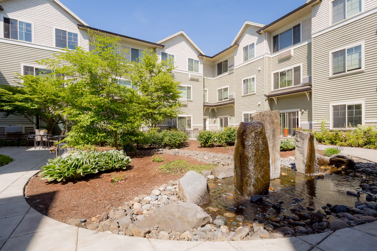Outdoor courtyard area of a senior living facility with a small water feature consisting of three vertical stone fountains surrounded by rocks. The courtyard is landscaped with green bushes, trees, and plants, and is enclosed by a three-story beige building with multiple windows. There are paved walkways and outdoor seating areas visible.