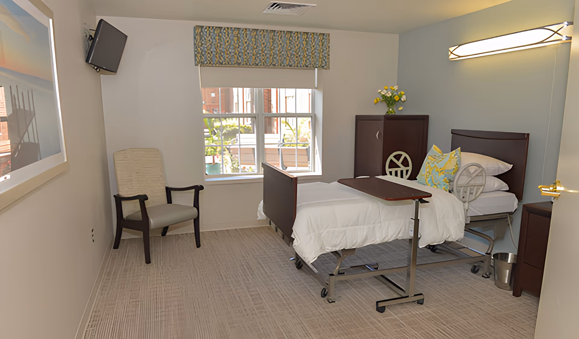 A bright single-occupancy senior living bedroom with a hospital-style bed, overbed table, armchair, window, and cabinet.