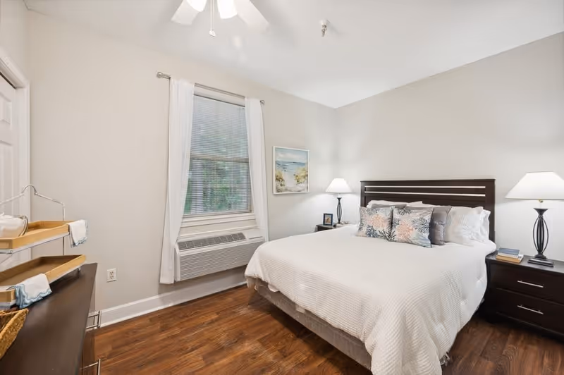 A bright and clean bedroom with a large bed covered in white bedding and decorative pillows. The room has wooden flooring, a window with white curtains, two bedside tables each with a lamp, a dresser with towels, and a ceiling fan. A framed picture hangs on the wall beside the bed.