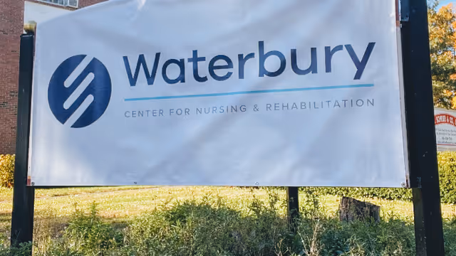 A large white sign with the logo and text for Waterbury Center for Nursing & Rehabilitation, mounted on black posts with grass and bushes in the foreground and a brick building partially visible in the background.