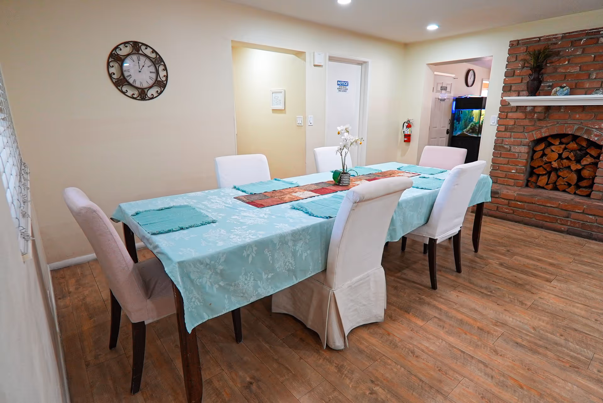 A dining room with a long table covered by a light blue tablecloth surrounded by upholstered chairs and a brick fireplace with stacked firewood.