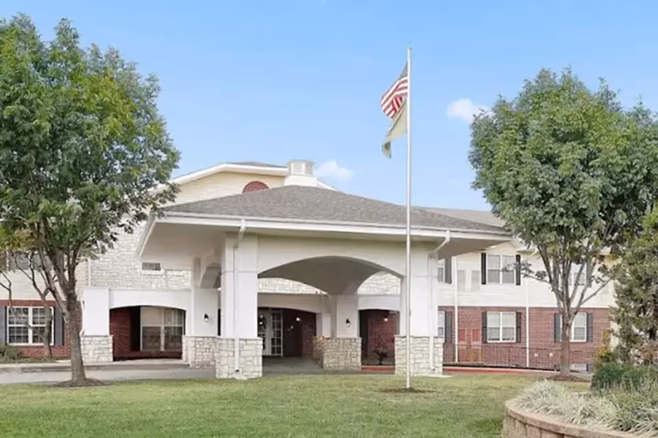 Front exterior of a senior living facility with a covered entrance, flagpole, and trees on a grassy lawn.