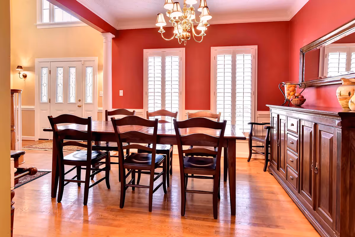 A dining room with a wooden table and six matching chairs on a hardwood floor. The walls are painted red with white trim, and there are two large windows with white shutters. A wooden sideboard with decorative items and a large mirror above it is on the right side. A chandelier with multiple lights hangs from the ceiling. The entrance door and a staircase are visible in the background.