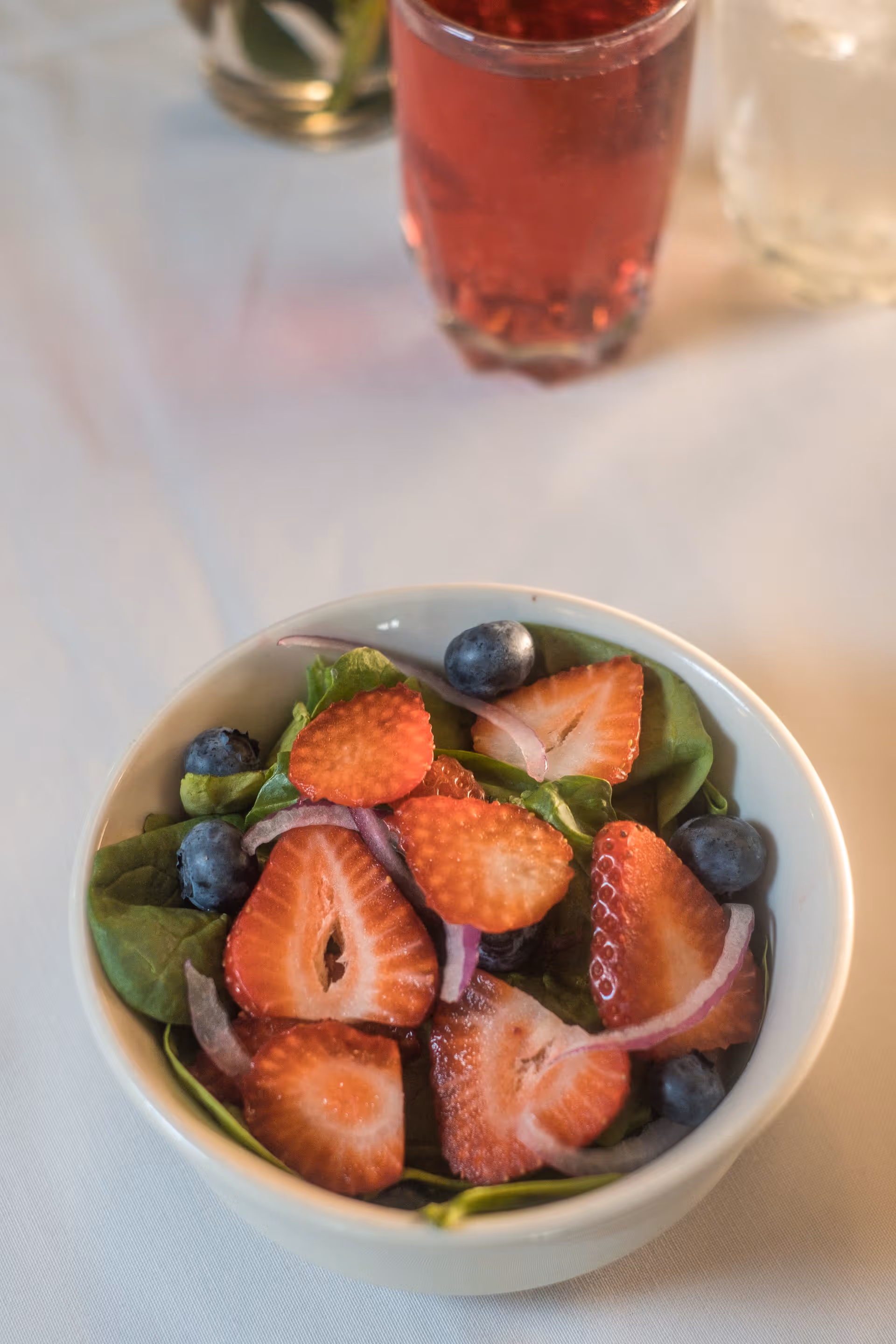 A bowl of fresh salad containing spinach leaves, sliced strawberries, blueberries, and thin slices of red onion on a white tablecloth with two glasses of beverages in the background.