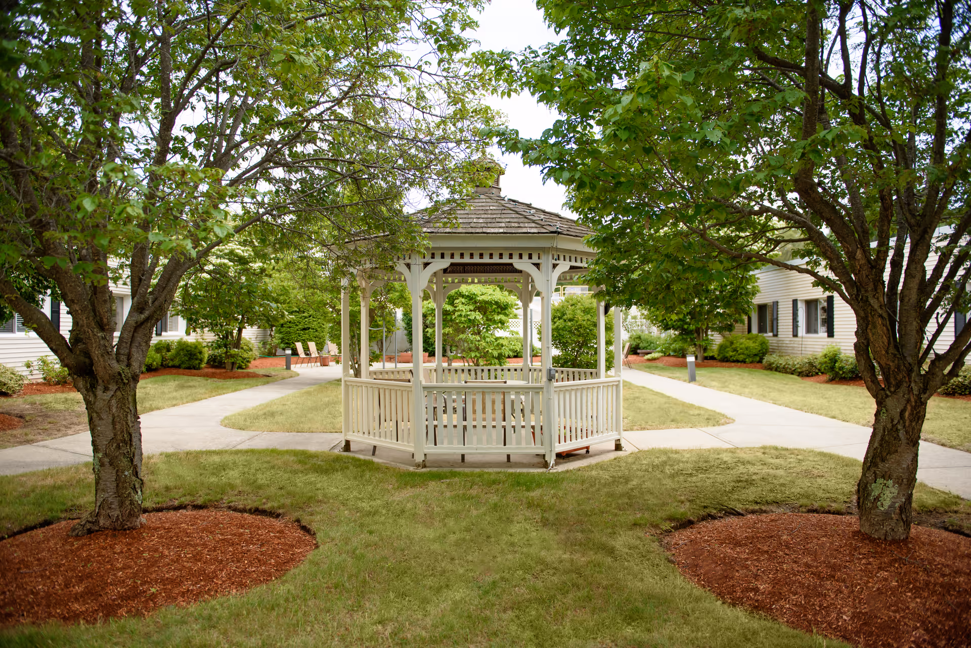 White wooden gazebo centered in a courtyard with pathways, trees, and surrounding senior living buildings.