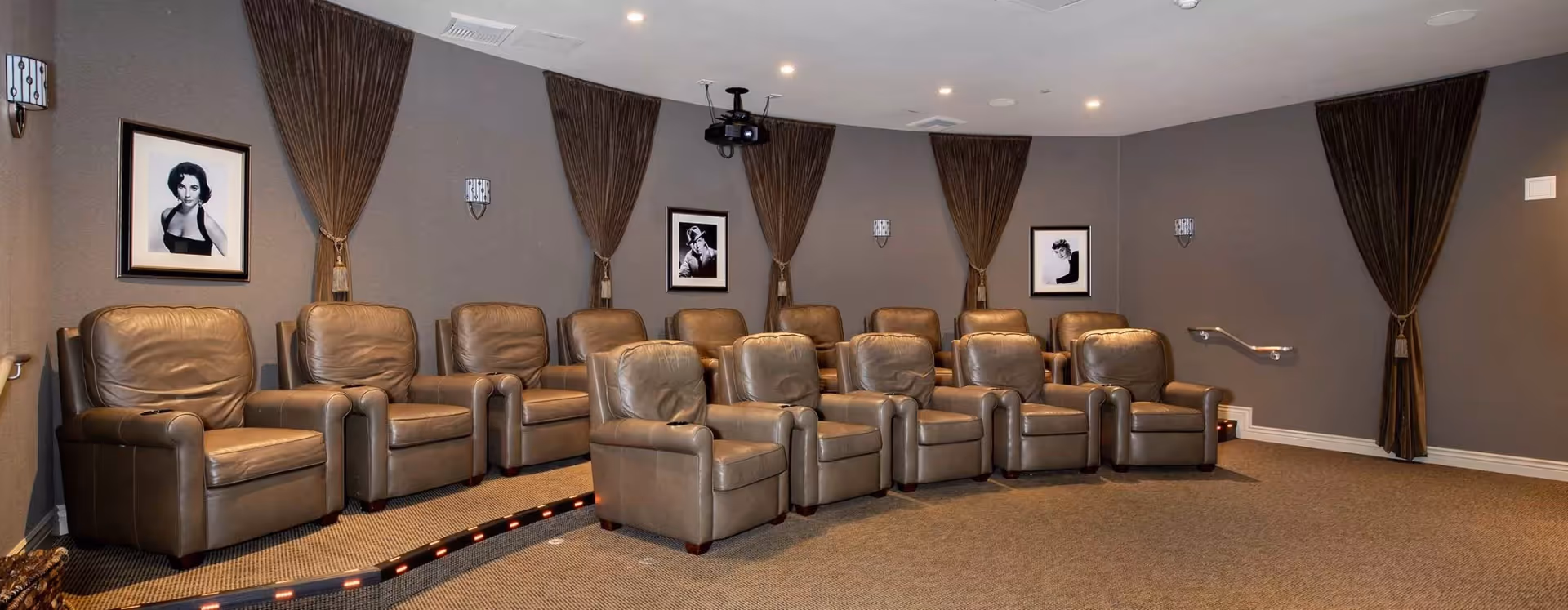 A small theater room with two rows of brown leather recliner chairs arranged on a carpeted floor. The walls are painted gray and decorated with black and white framed portraits and brown curtains tied back with tassels. Ceiling lights illuminate the room and a projector is mounted on the ceiling.