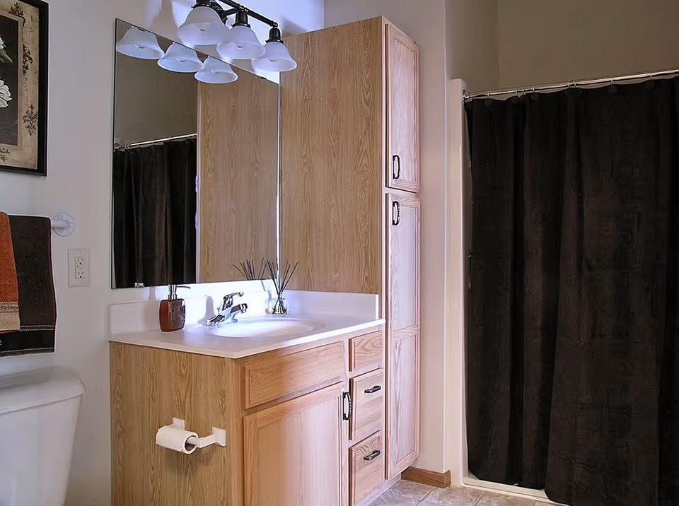 Bathroom with a wooden vanity cabinet, white countertop with a sink, a large mirror above the sink, a light fixture with five lamps, a toilet with a toilet paper holder, a towel rack with folded towels, and a shower area with a dark curtain.