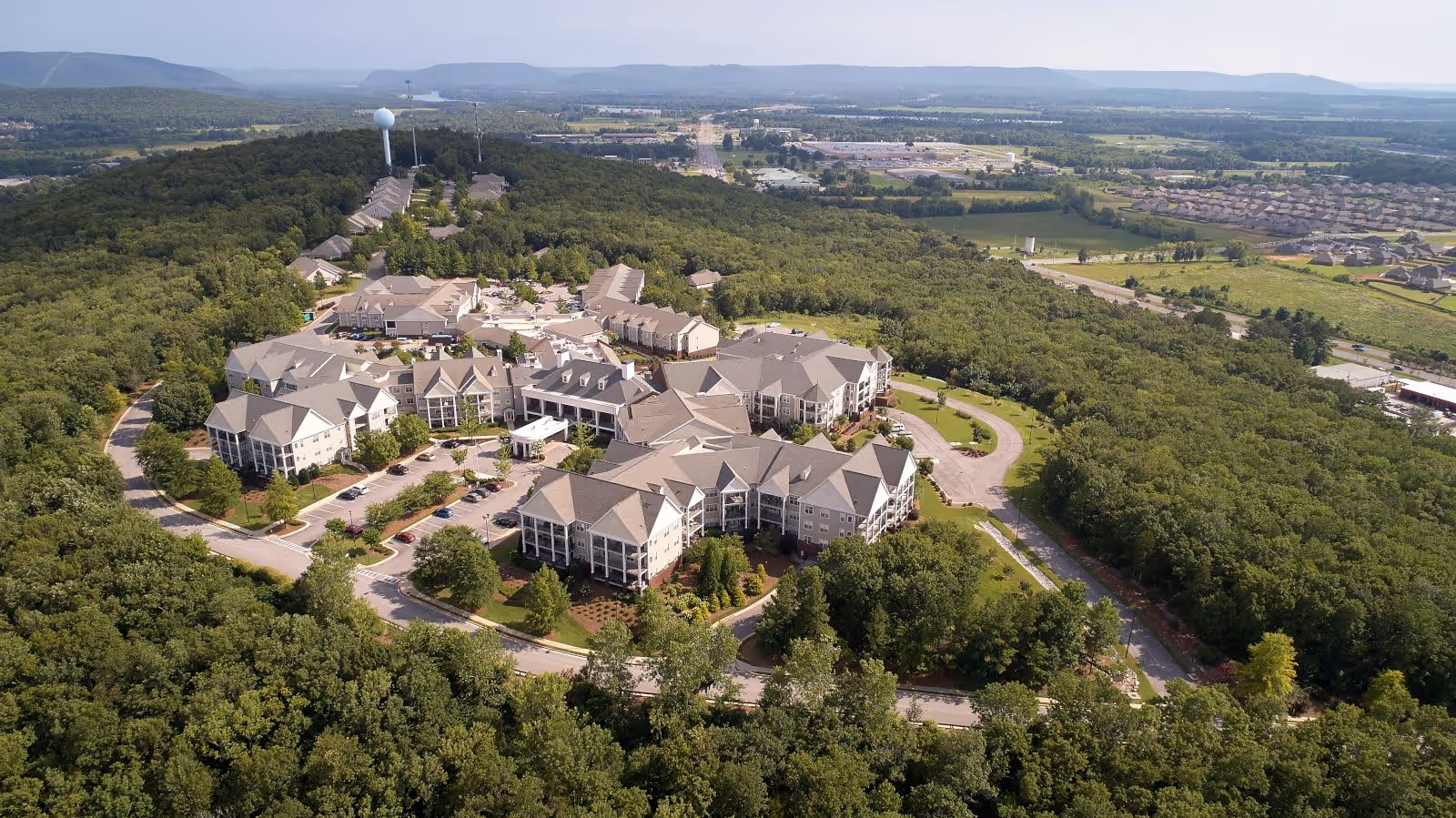 Aerial view of Redstone Village senior living facility surrounded by dense green forest with multiple buildings and parking areas. The landscape extends to distant hills and residential areas under a clear sky.