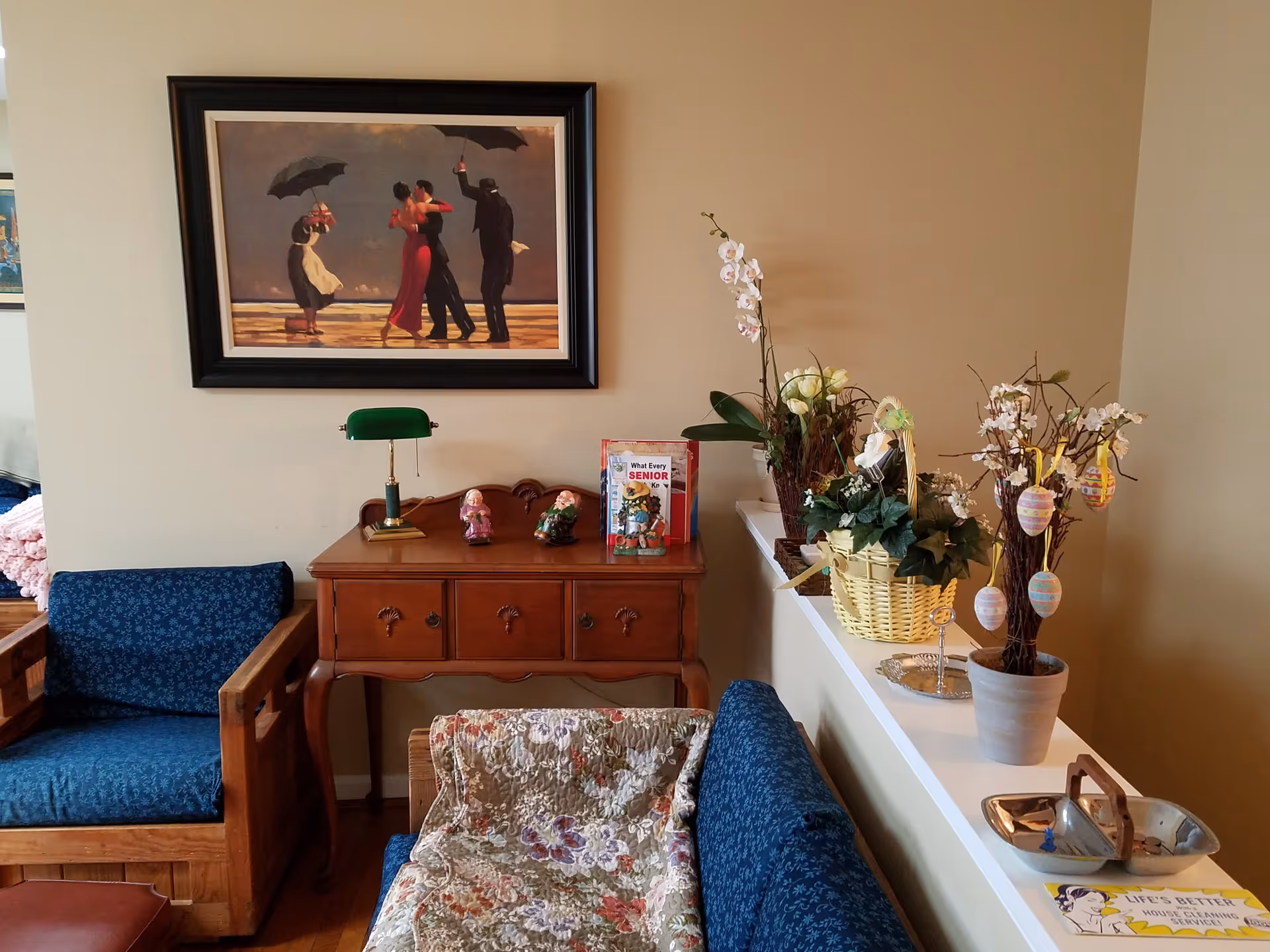 A cozy living room area with wooden framed blue upholstered chairs and a floral quilt draped over one chair. A wooden side table with drawers holds a green banker’s lamp, two small figurines, and a book titled 'What Every Senior'. Above the table hangs a framed painting of four people dancing on a beach, two holding umbrellas. To the right, a white ledge displays several flower arrangements and a basket with decorative branches holding pastel-colored Easter eggs. The walls are painted beige.