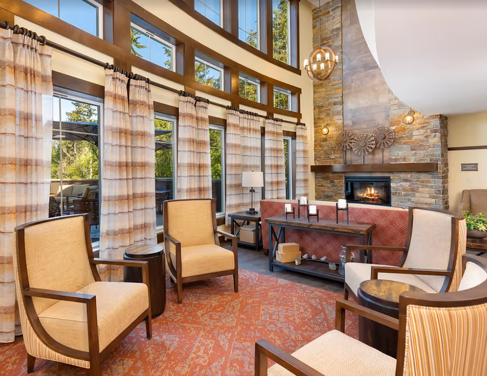 Bright communal living room with tall windows and curtains, a stone fireplace, and several armchairs arranged around tables.