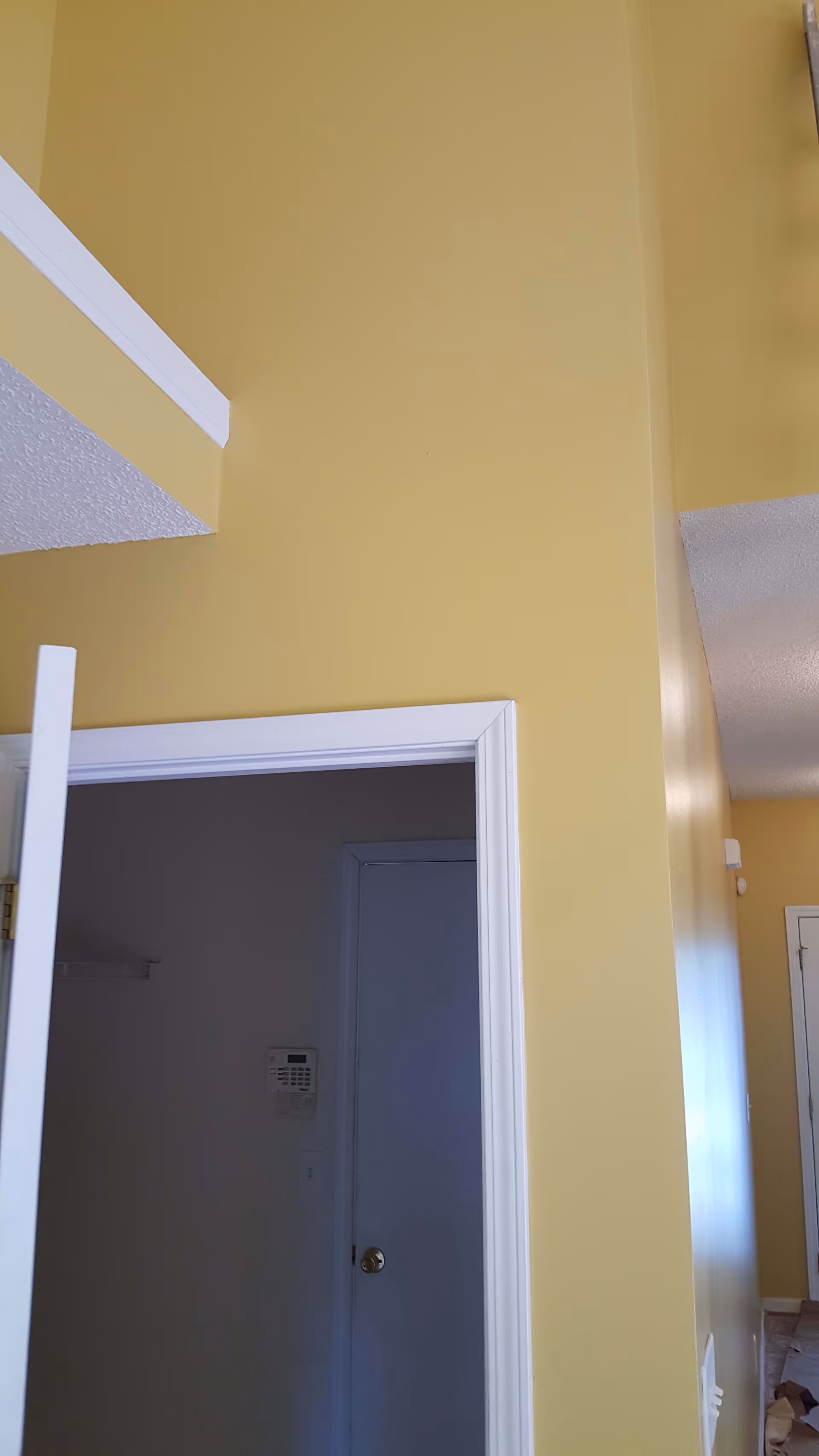 Interior view of a hallway with yellow walls and white door frames. One door is partially open, revealing a dark room with a security keypad on the wall. The ceiling has a textured finish.
