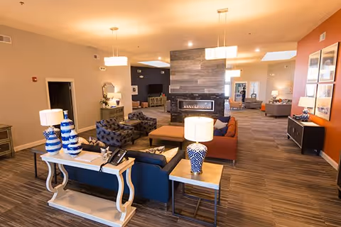 Spacious senior living lounge with sofas, armchairs, tables, lamps, and a central double-sided fireplace under warm lighting.