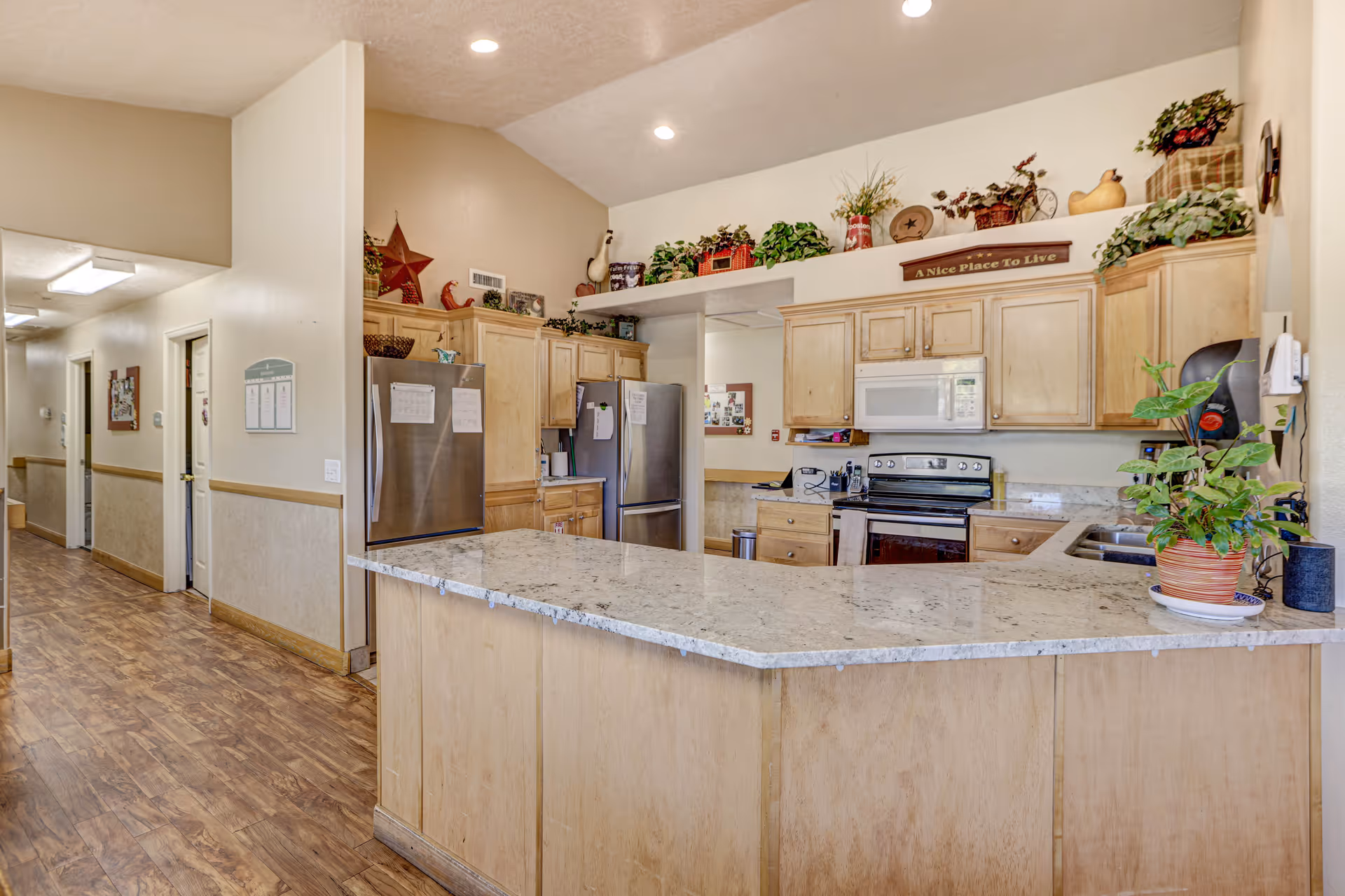 A spacious kitchen area with light wood cabinets, a large granite countertop island, stainless steel refrigerator, stove, and microwave. The kitchen is decorated with various plants and decorative items on top of the cabinets. To the left, there is a hallway with wooden flooring and beige walls leading to other rooms.