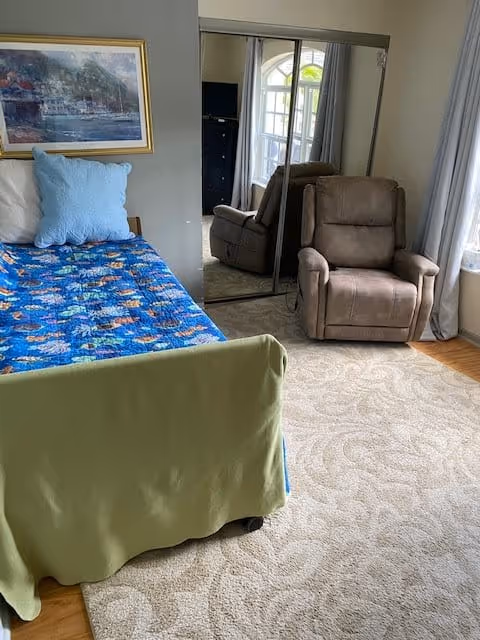A bedroom with a single bed covered with a blue blanket featuring a colorful pattern, a light blue pillow, and a green blanket draped over the foot of the bed. Next to the bed is a brown recliner chair on a beige patterned carpet. A large mirrored closet door reflects part of the room, and a window with light curtains allows natural light to enter. A framed painting hangs on the wall above the bed.