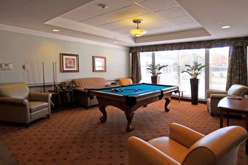 A carpeted communal lounge with a pool table surrounded by armchairs, a sofa, side tables and large windows.