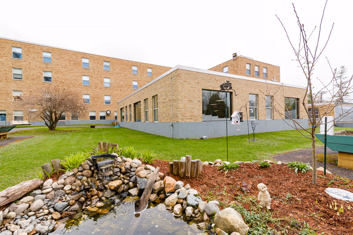 Outdoor garden area at St. Vincent de Paul Rehabilitation and Nursing Center featuring a small pond with rocks and a water feature, landscaped flower beds with mulch, a few small trees, and a brick building in the background.