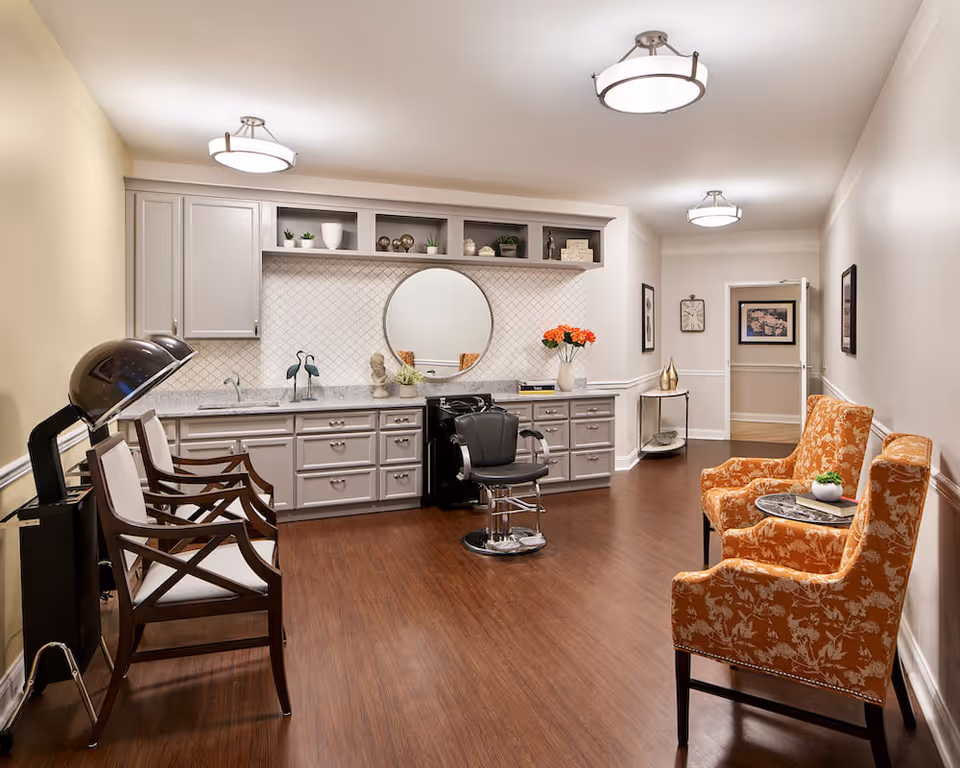 Bright salon-style interior with a styling chair and hooded dryer, long cabinetry and countertop, and orange patterned guest chairs in a waiting area.