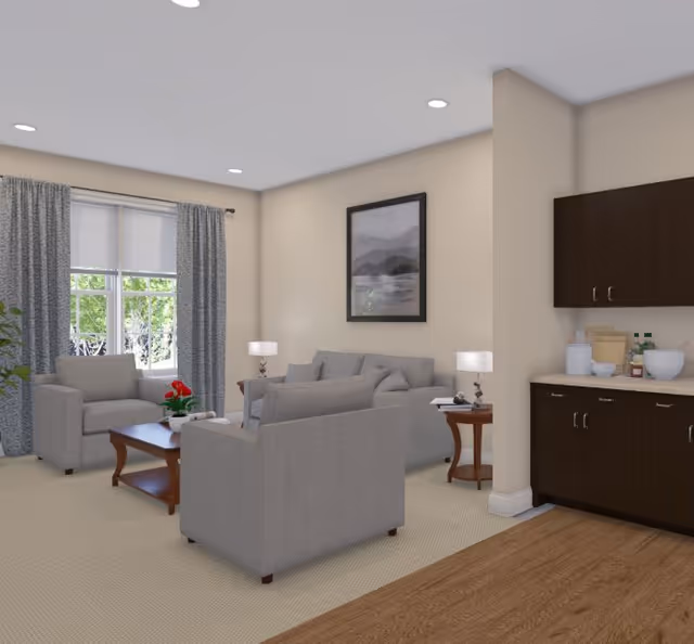 A cozy living room area with two gray armchairs and a gray sofa arranged around a wooden coffee table with a red flower centerpiece. The room has beige walls, a large window with gray curtains, two side tables with lamps, a framed landscape picture on the wall, and a kitchenette area with dark wood cabinets and a countertop with kitchen items.