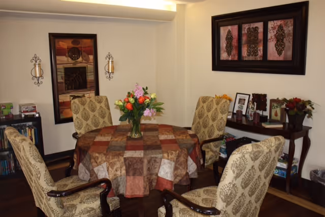 A cozy dining area with a round table covered by a patchwork tablecloth in shades of brown, beige, and rust. Four upholstered chairs with patterned fabric surround the table. A vase with colorful flowers is placed in the center of the table. The room has light-colored walls adorned with framed artwork and decorative wall sconces. A small bookshelf and a console table with framed photos and decorative items are visible in the background.