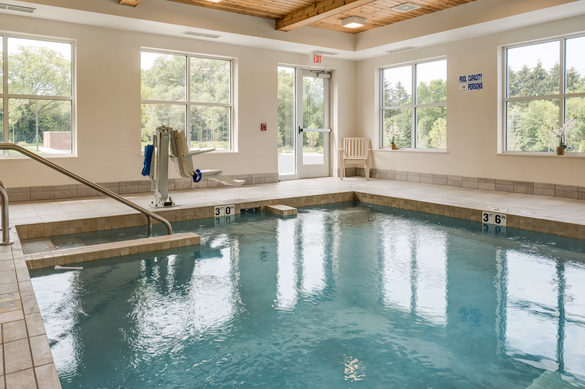 Indoor swimming pool with clear blue water, surrounded by tiled flooring and large windows showing green trees outside. There is a pool lift chair for accessibility, a metal handrail for entering the pool, and a white chair near the exit door. The ceiling has wooden beams and recessed lighting.
