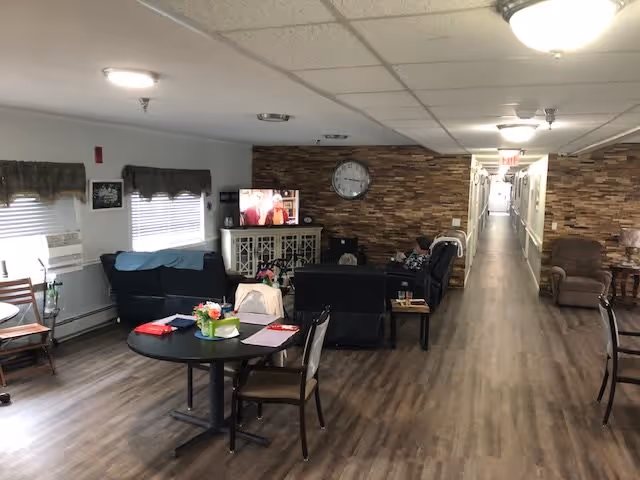 Interior view of a senior living facility common area with a round table and chairs in the foreground, a seating area with sofas and recliners facing a TV mounted on a cabinet, a clock on a stone accent wall, and a long hallway extending into the distance.