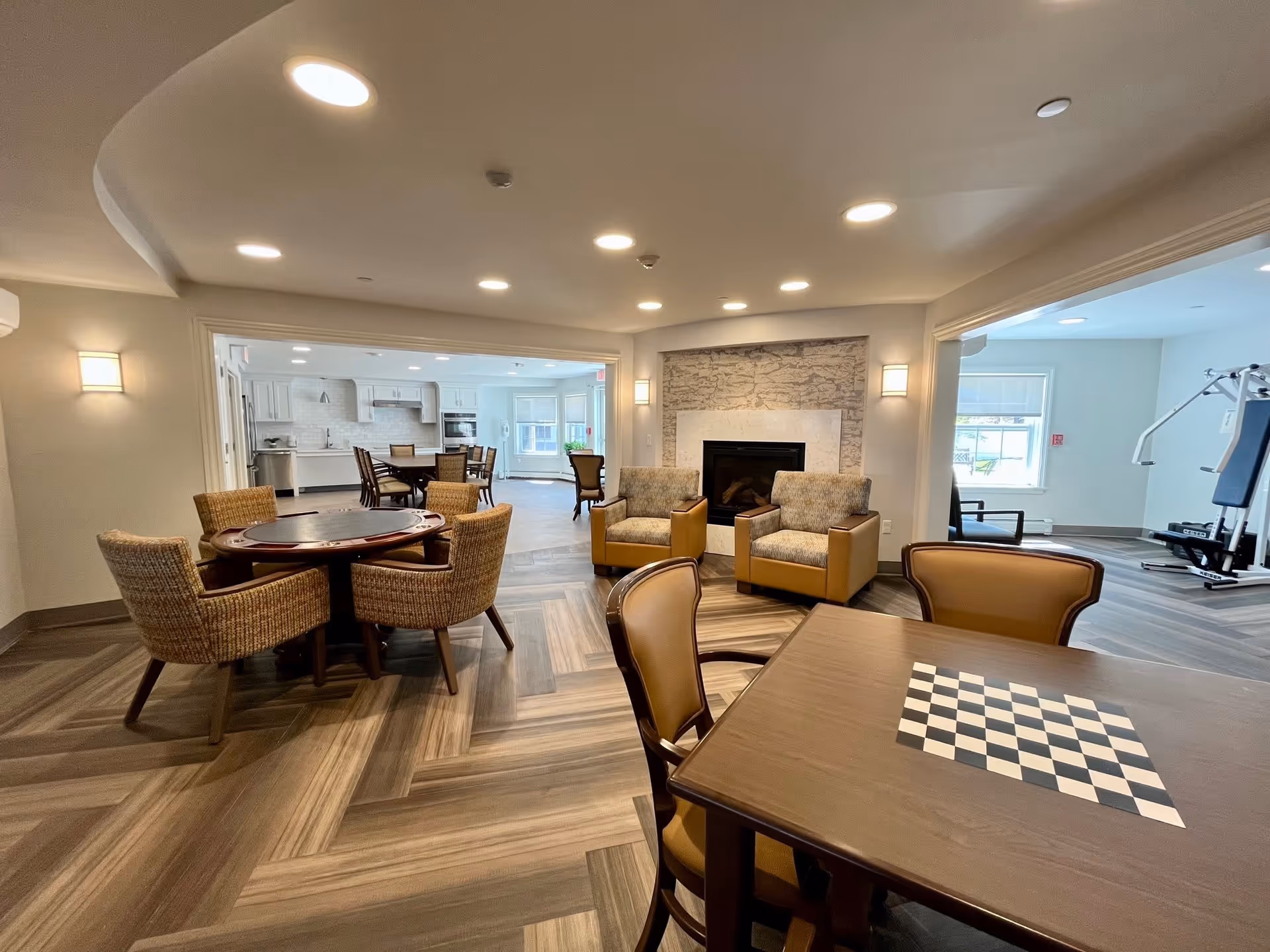 Community lounge with multiple game tables, upholstered chairs, a fireplace, and an open view into a kitchen and fitness area.