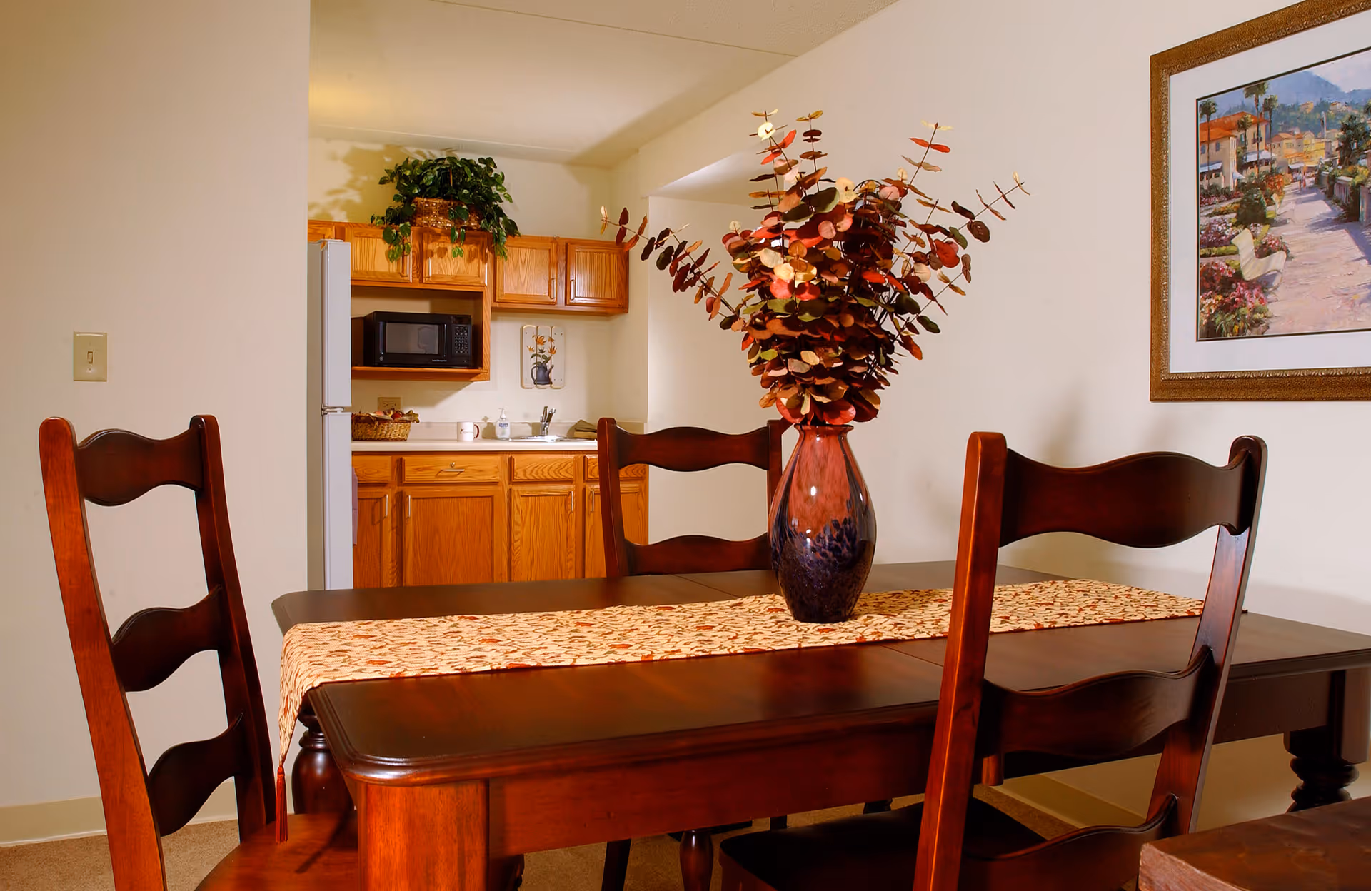 Dining area with a dark wooden table and four matching chairs. A decorative vase with red and brown foliage sits on a patterned table runner. In the background, a small kitchen area with wooden cabinets, a microwave, a refrigerator, and a sink is visible. A framed painting hangs on the wall to the right.