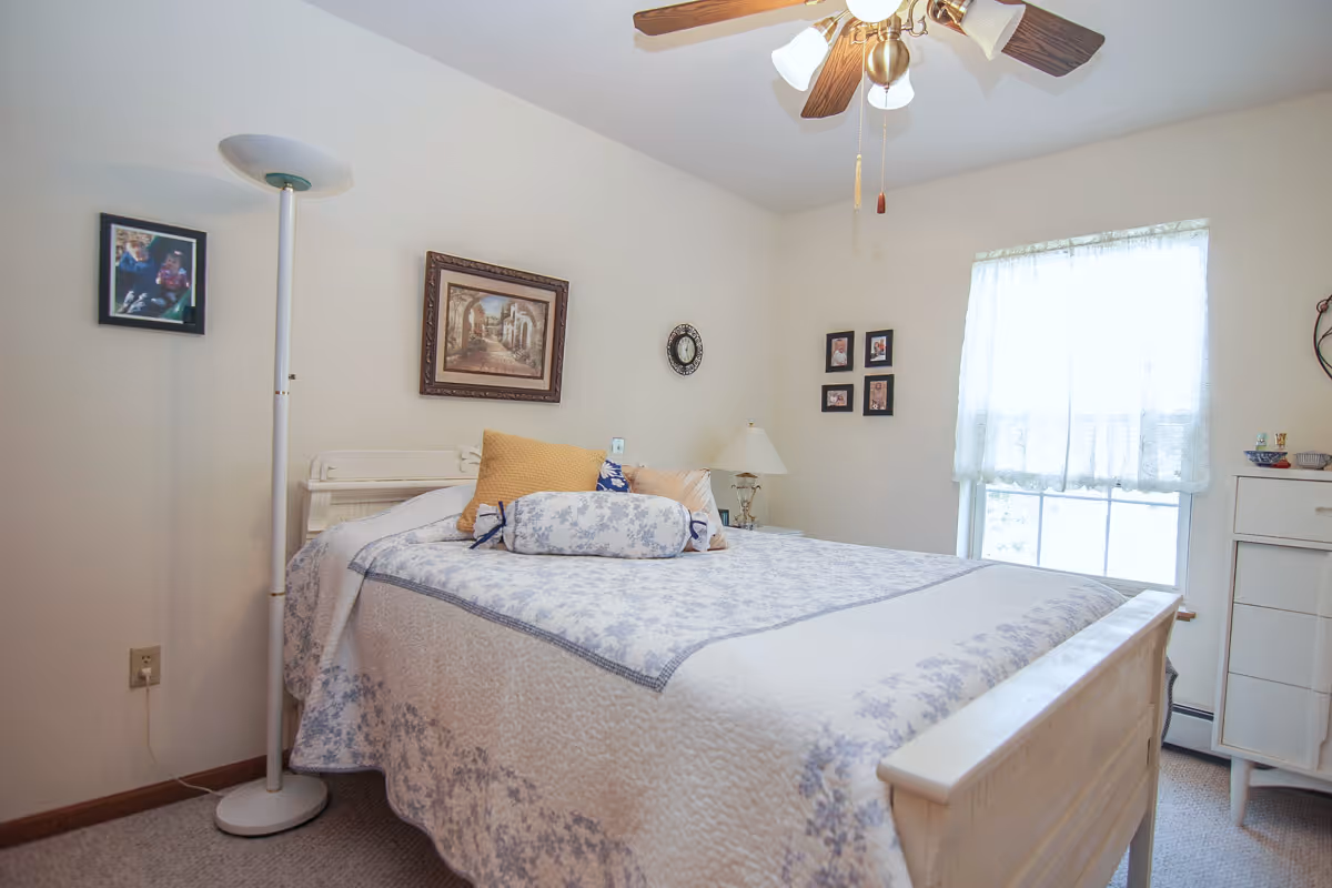 Bright bedroom with a bed covered in a patterned quilt and pillows, a floor lamp, wall art, ceiling fan, and a window with a lace curtain.