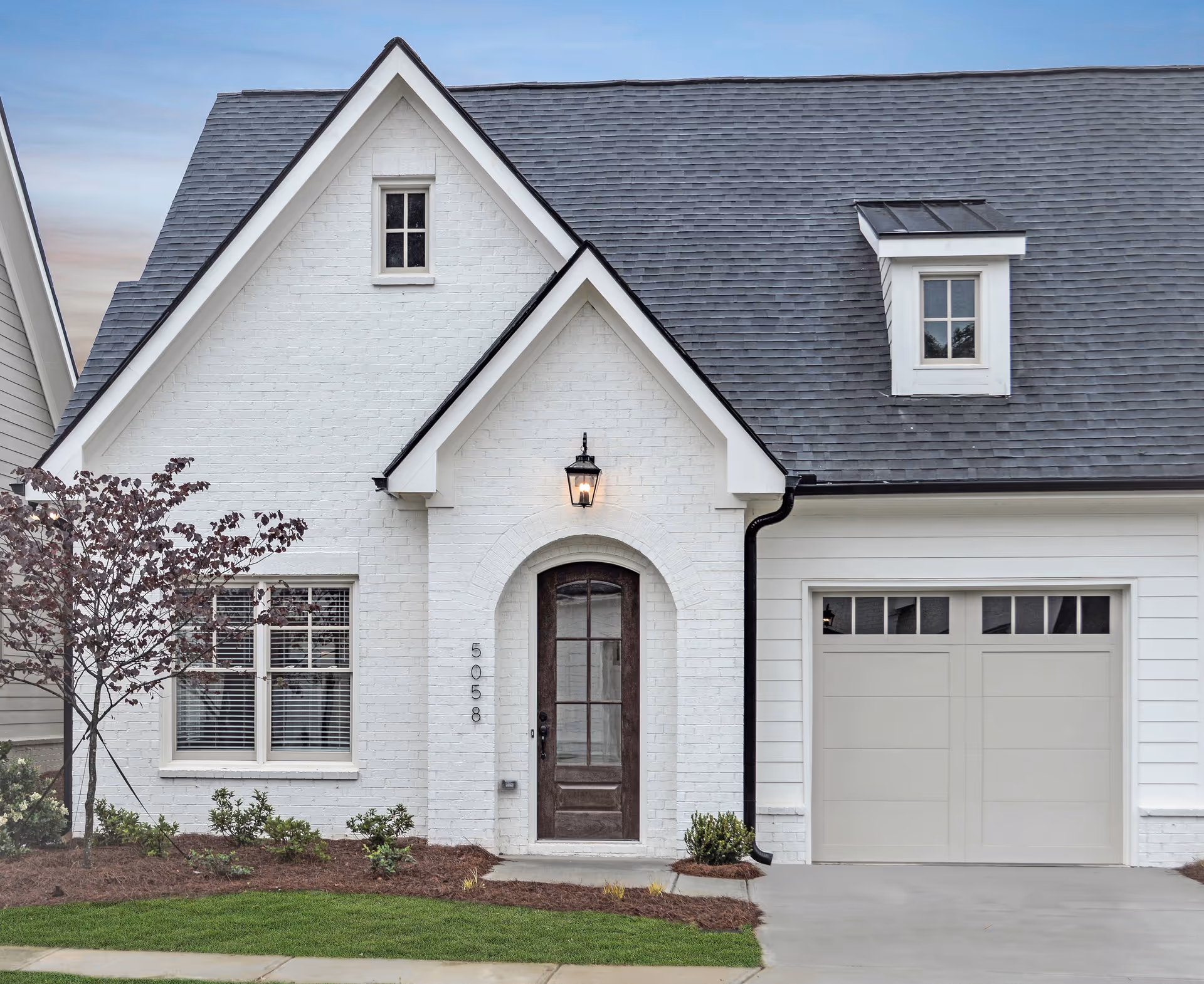 Front exterior view of a white brick house with a dark wooden front door, a small window above the door, a garage with a light gray door, and a small tree and shrubs in the front yard.