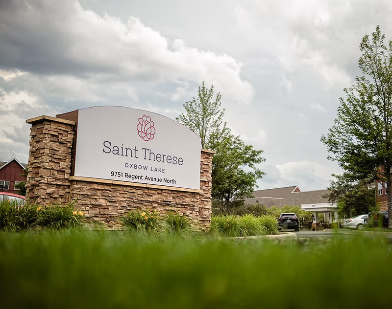 Stone monument sign for Saint Therese Oxbow Lake with address 9751 Regent Avenue North, surrounded by greenery and trees, with a cloudy sky and part of the senior living facility visible in the background.