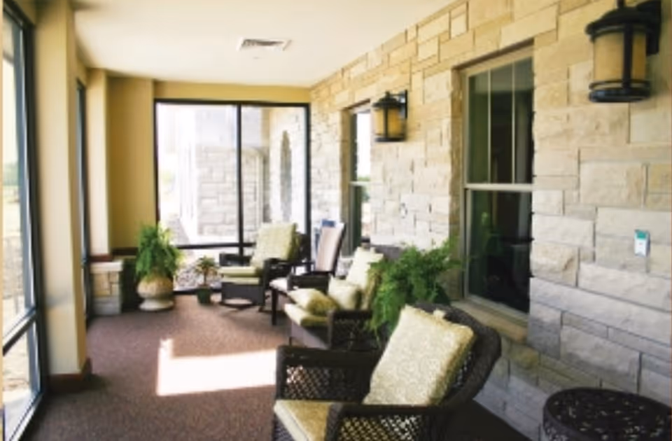 A sunlit indoor seating area with cushioned wicker chairs and potted plants along a stone wall with windows and wall-mounted lanterns.