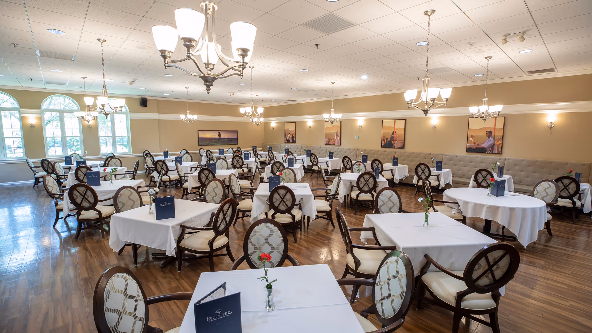 A spacious dining room with multiple tables covered in white tablecloths, each set with a small vase holding a single flower and a menu card. The room features wooden floors, elegant chandeliers, large windows letting in natural light, and framed pictures on the walls.