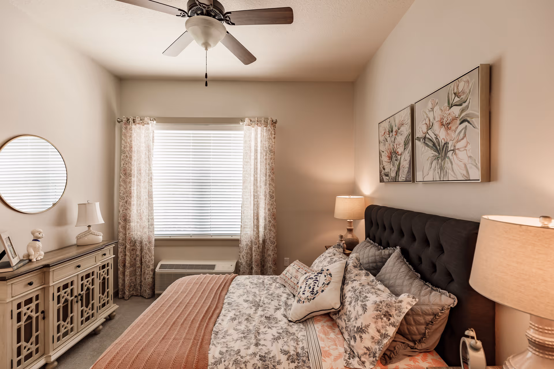 A cozy bedroom with a large bed featuring floral and patterned bedding, multiple pillows, and a tufted dark headboard. On the right side of the bed is a nightstand with a lamp and an alarm clock. Above the bed are two framed floral paintings. Across from the bed is a window with blinds and patterned curtains, and below the window is an air conditioning unit. To the left of the window is a decorative cabinet with a round mirror above it and a small lamp and figurine on top. A ceiling fan with wooden blades is mounted on the ceiling.