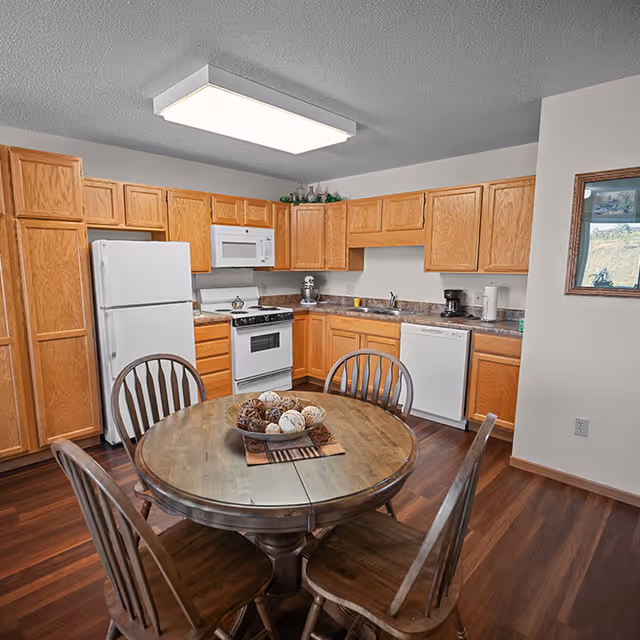 A kitchen with wooden cabinets, a white refrigerator, stove, microwave, dishwasher, and a round wooden dining table with four chairs. The table has a decorative centerpiece with assorted decorative balls. The floor is wooden, and there is a framed picture on the wall.